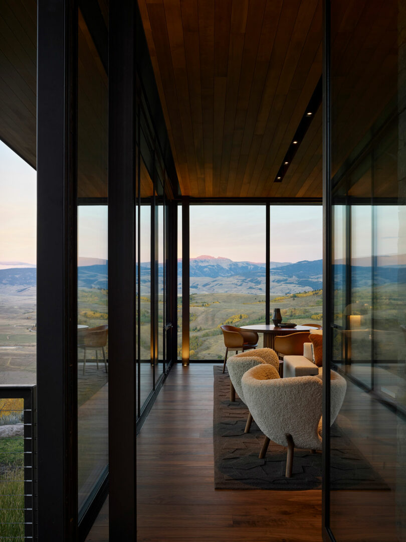 Una moderna habitación con paredes de vidrio con dos sillones y una pequeña mesa domina un pintoresco paisaje de montaña al atardecer.