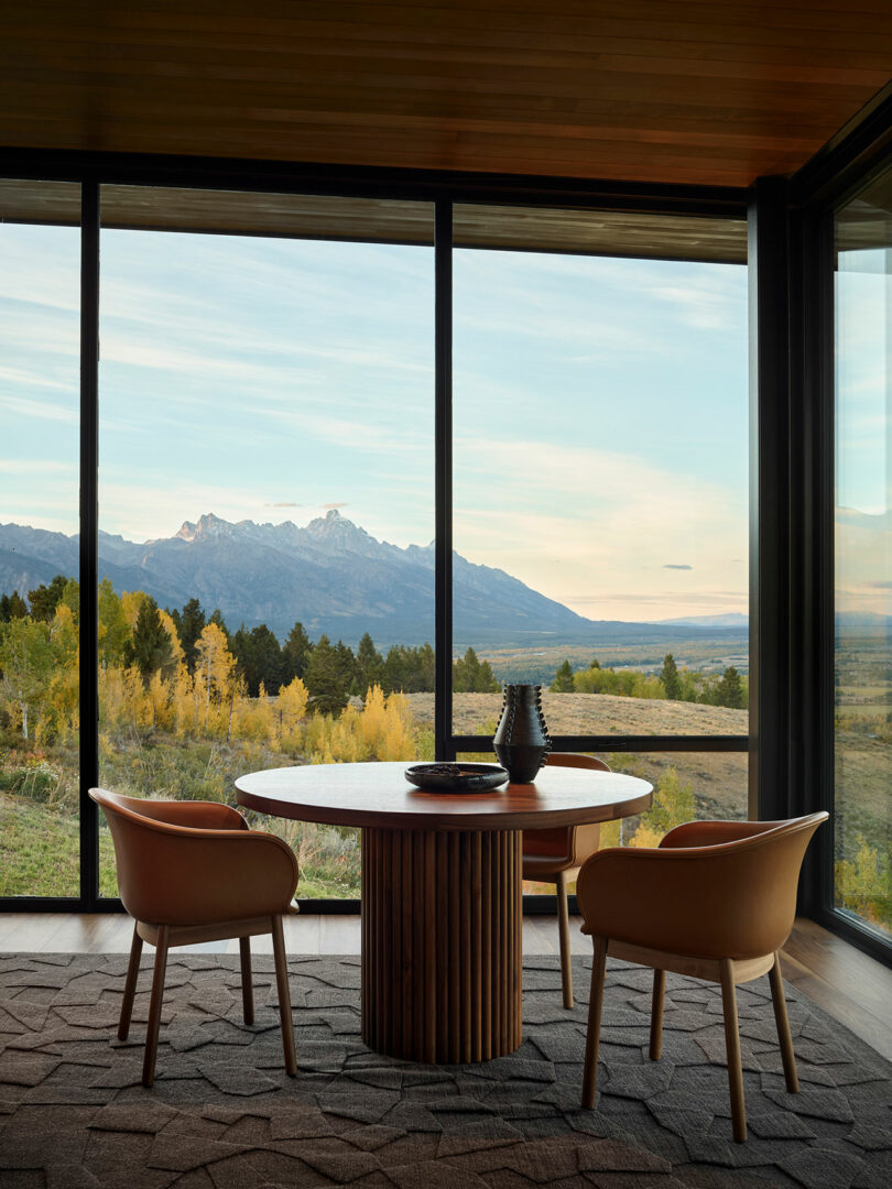Una mesa redonda de madera con dos sillas se encuentra en grandes ventanas de piso a techo con vistas a un paisaje pintoresco de montañas, árboles y cielo.