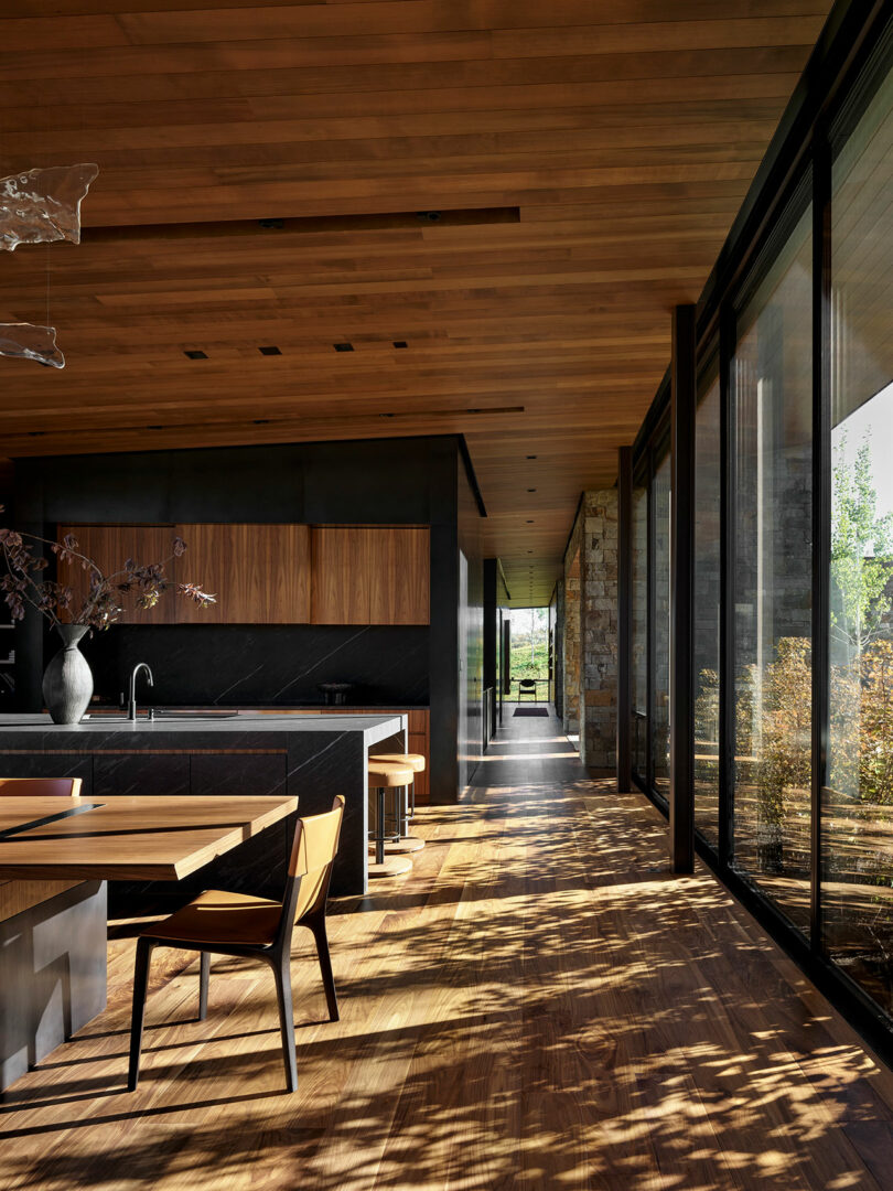Cocina moderna y comedor con pisos de madera, techos de madera, gabinetes oscuros, ventanas grandes y luz de sol que arrojan sombras desde árboles fuera del espacio interior.