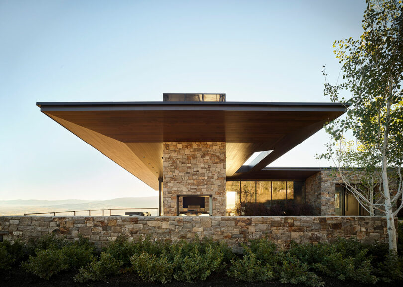 Casa moderna con techo de madera ancho y amplio, paredes de piedra, grandes ventanas y una chimenea al aire libre, ubicada contra un paisaje natural y un cielo claro.