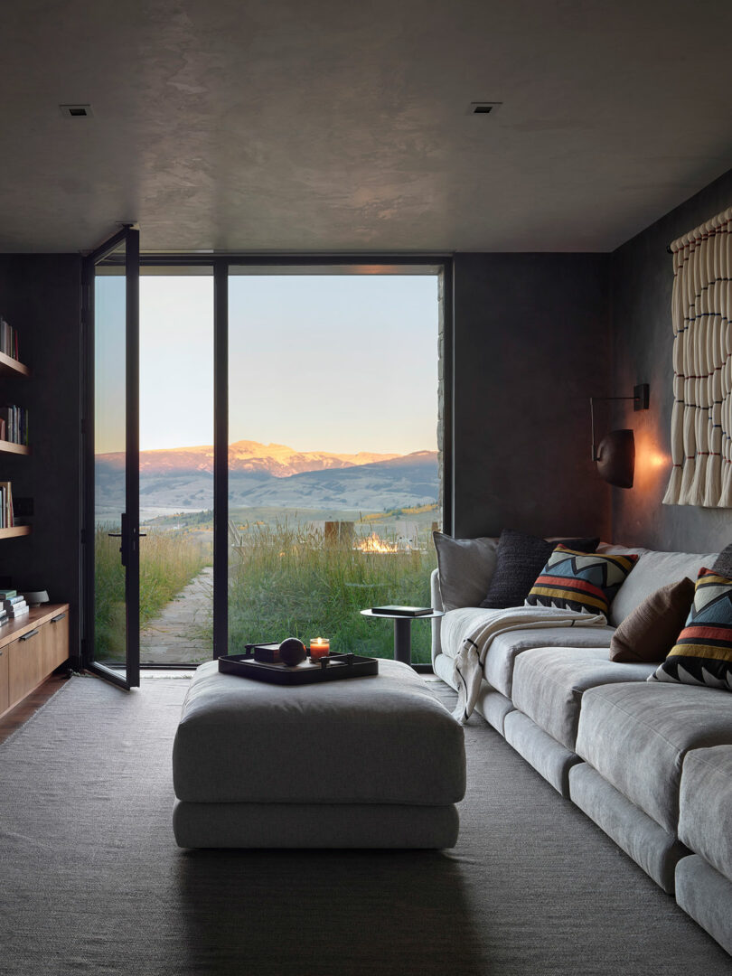 Una sala de estar moderna con un sofá seccional gris, una gran otomana y una puerta de vidrio abierta que revela un pintoresco paisaje de montaña al atardecer.