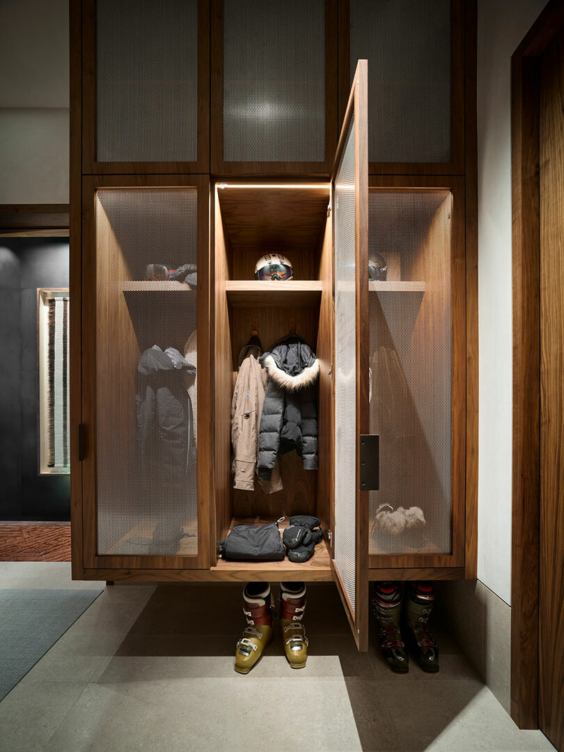 Locker de madera con puertas de malla, que contienen abrigos de invierno, cascos, guantes y botas de esquí cuidadosamente dispuestas en estantes y el piso.