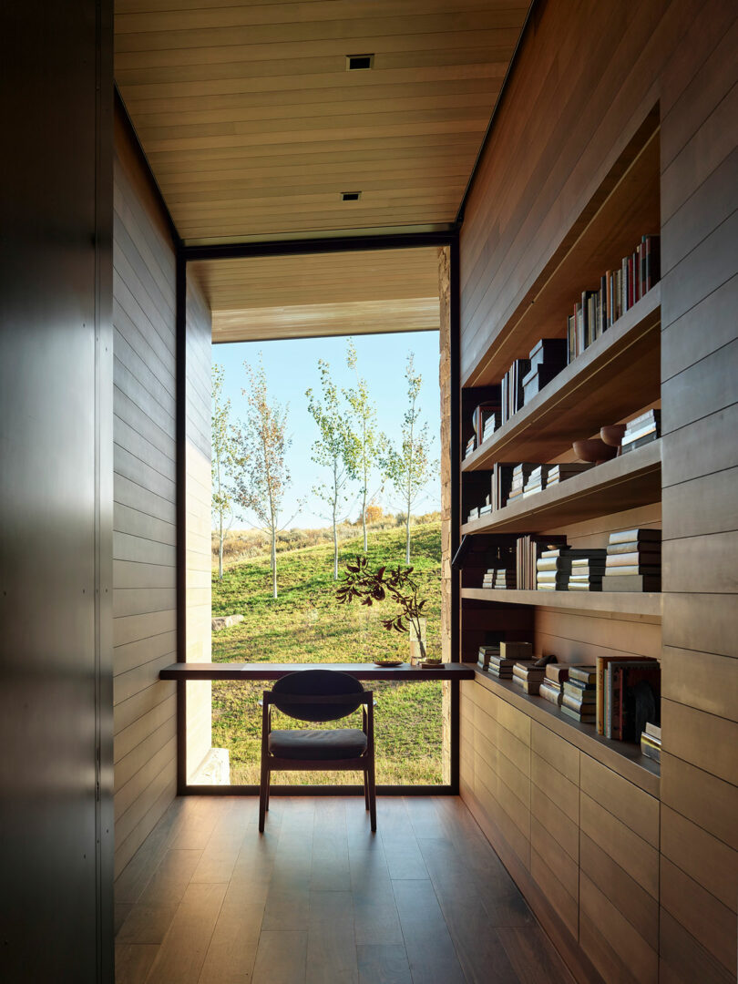 Un estudio moderno estrecho con paredes con paneles de madera, estanterías incorporadas, un escritorio y una silla, con una gran ventana con vistas a un paisaje cubierto de hierba con árboles.