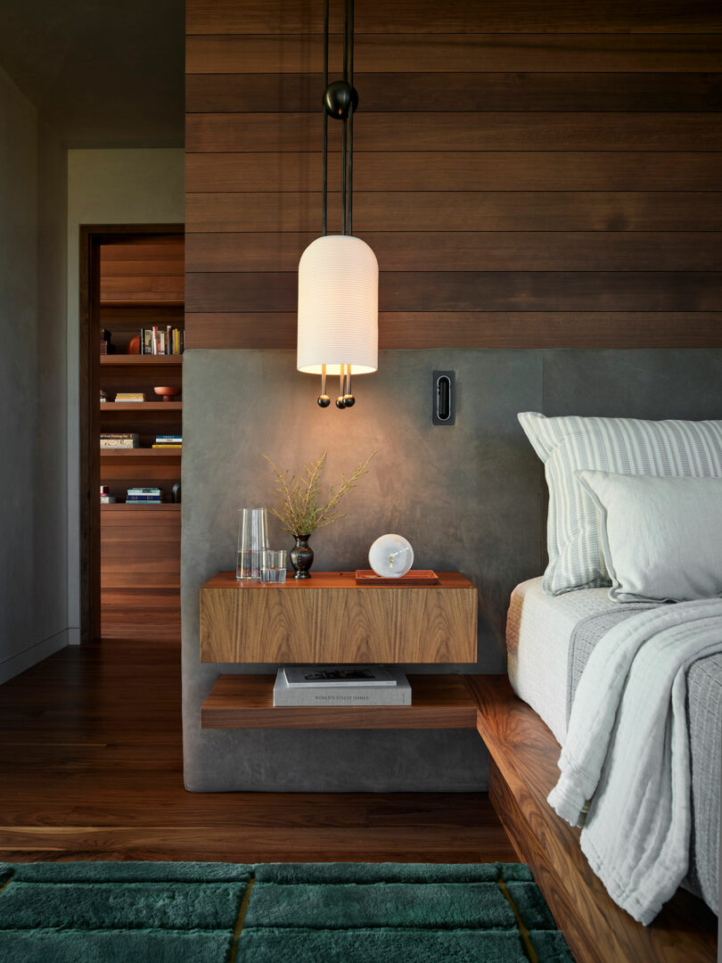 Un dormitorio moderno con una pared de acento de madera, luz colgante, mesita de noche flotante que sostiene un jarrón, vidrio y reloj, y una cama con ropa de cama a rayas. Una estantería es visible en el fondo.