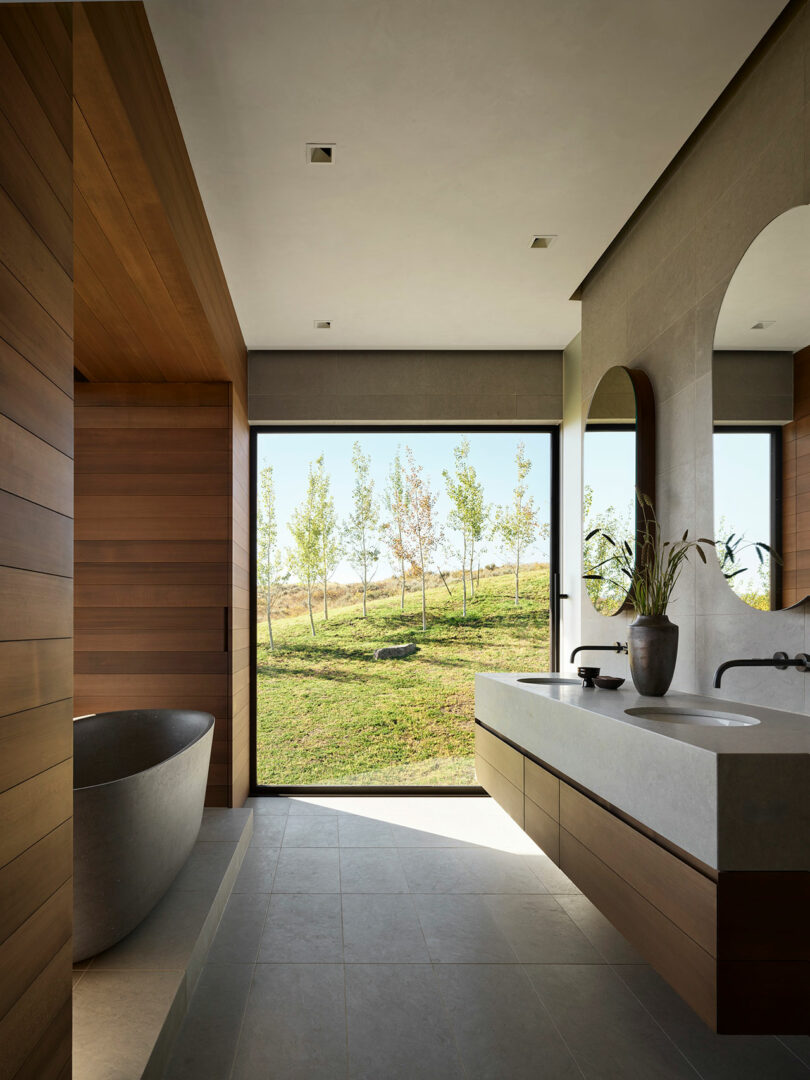 Un baño moderno con madera y acabados de piedra, una bañera independiente, un tocador grande y una ventana de piso a techo con vistas a un paisaje de hierba al aire libre.