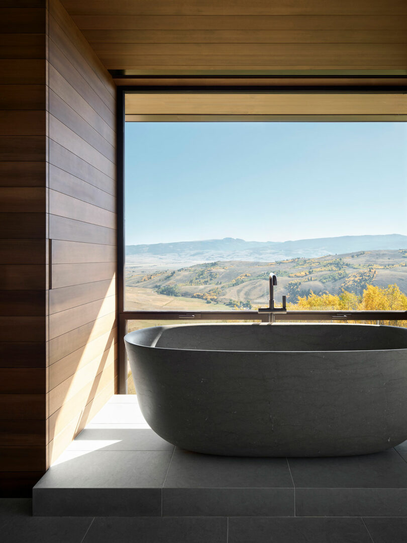 Una moderna bañera negra independiente está colocada por una gran ventana con una pintoresca vista de montaña y valle; La habitación cuenta con superficies de madera y baldosas.