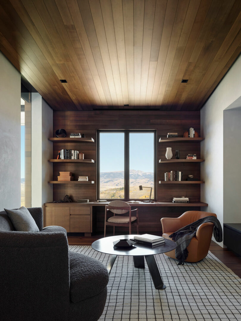Moderna oficina en casa con techo de madera y estantes, un escritorio que mira una ventana con una vista de montaña, un sofá gris, un sillón marrón y una mesa de café redonda en una alfombra estampada.