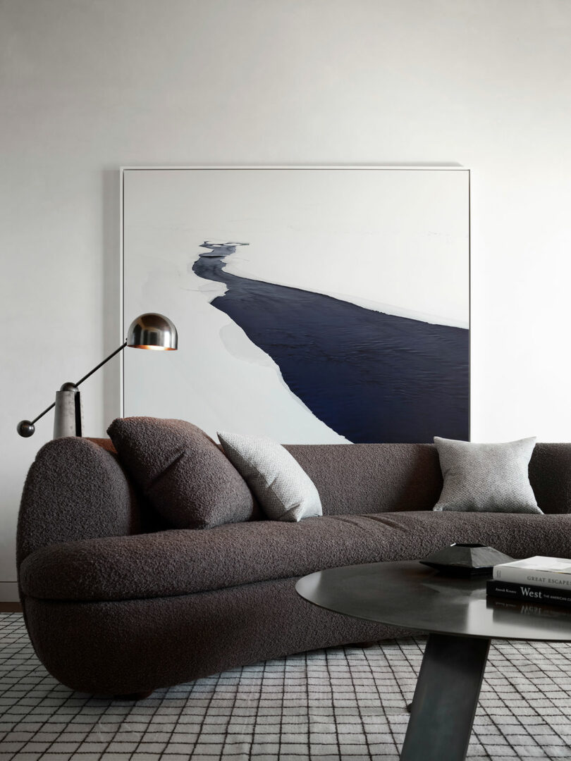 Una sala de estar moderna con un sofá de textura oscura, cojines monocromáticos, mesa de café negro redondo y una gran pintura abstracta con tonos azul marino y blanco en la pared.