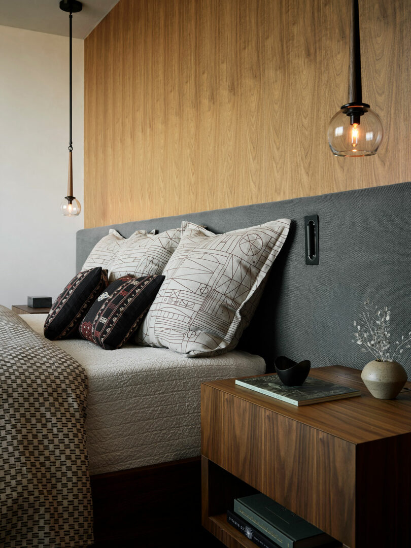 Una habitación moderna con cabecera tapizada gris, almohadas estampadas, una mesita de noche de madera con libros y decoración, y dos luces colgantes de vidrio colgante.