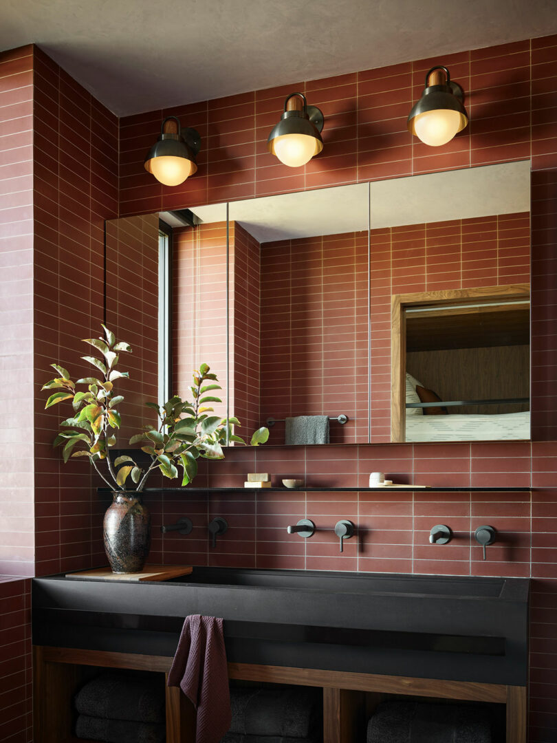 Baño moderno con paredes de azulejos rojos, una encimera negra, fregadero redondo, un espejo grande, tres luces montadas en la pared y un jarrón con ramas en el mostrador.