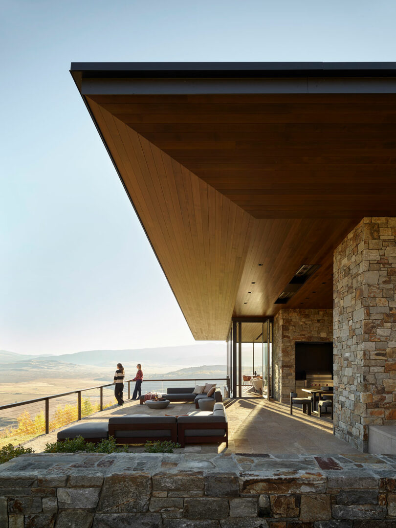 Casa moderna con paredes de piedra y un gran voladizo de madera, área de asientos al aire libre y dos personas paradas en una terraza con vistas a un paisaje pintoresco.