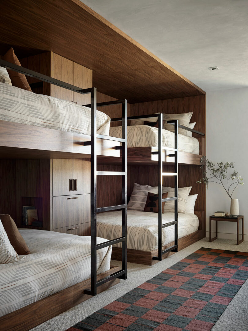 Cuatro literas de madera con escaleras de metal están dispuestas contra una pared en una habitación moderna; También se ve una mesa auxiliar con un jarrón y una alfombra geométrica.