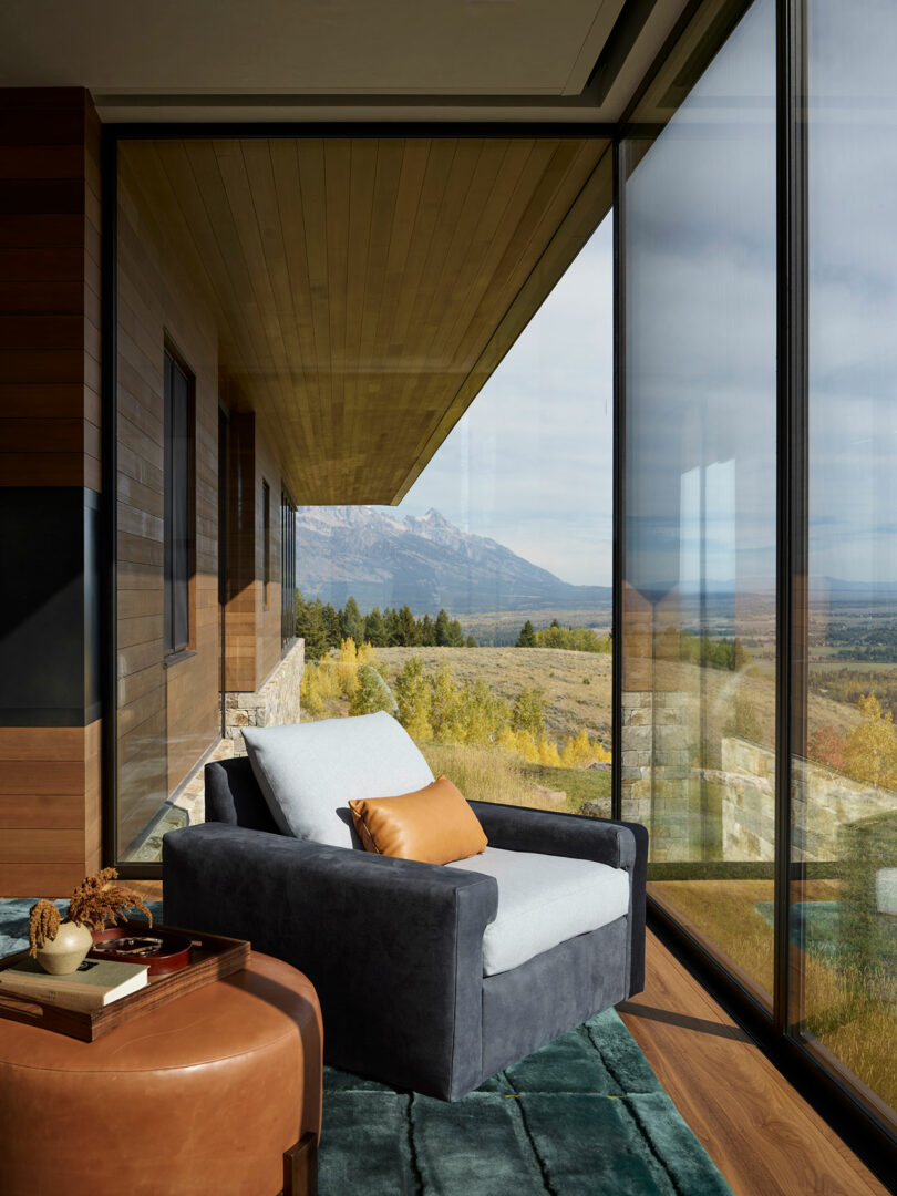 Un sillón moderno con una almohada bronceada se encuentra en ventanas de piso a techo, con vistas a un pintoresco paisaje de montaña y bosque.