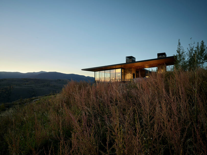 Casa moderna con grandes vidrieras situadas en una ladera cubierta de hierba al anochecer, con vistas a las montañas distantes bajo un cielo claro.