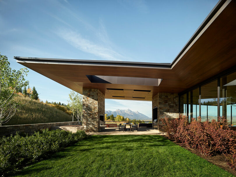 Una casa moderna con un techo amplio, paredes de piedra y vidrio, un patio al aire libre cubierto y césped ajardinado, con montañas visibles en el fondo.
