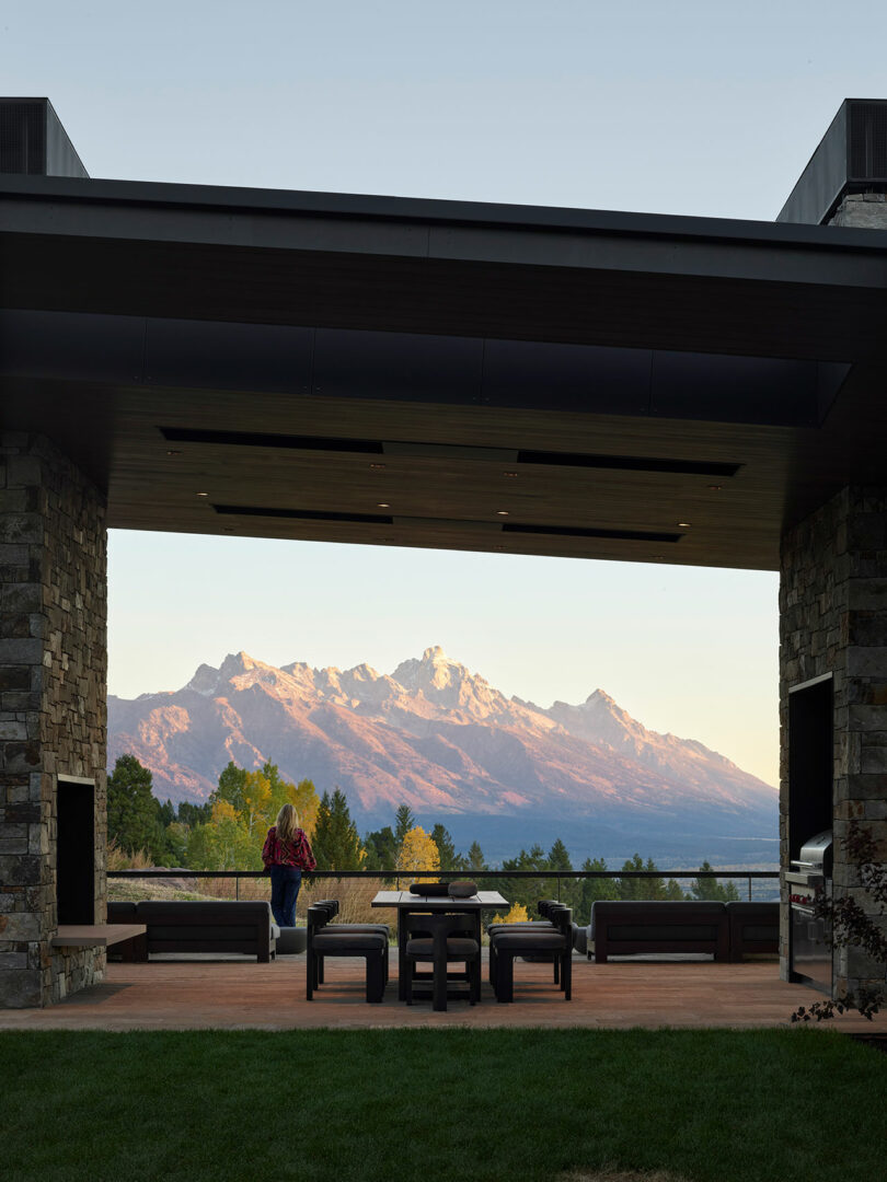 Una persona se sienta en un patio debajo de un área cubierta, frente a los muebles de exterior y una vista escénica de las montañas nevadas al atardecer.