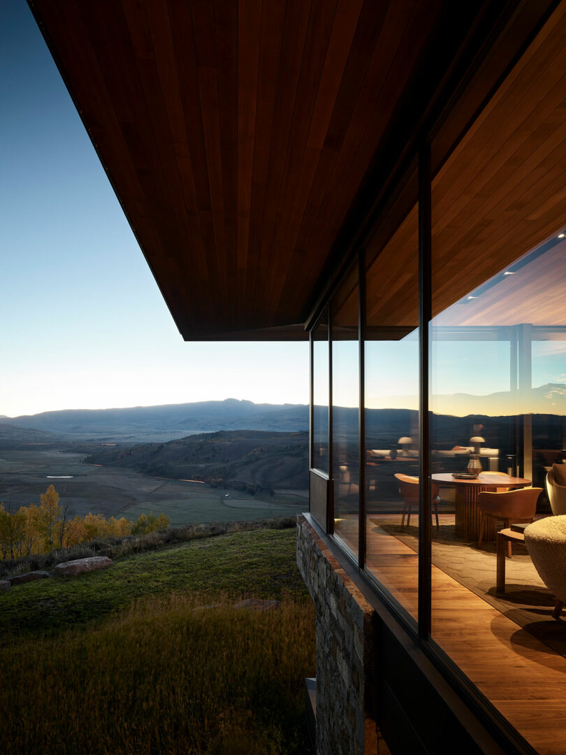 La casa moderna con grandes vidrios domina un valle pintoresco y montañas distantes al atardecer, con una cálida iluminación interior visible en el interior.