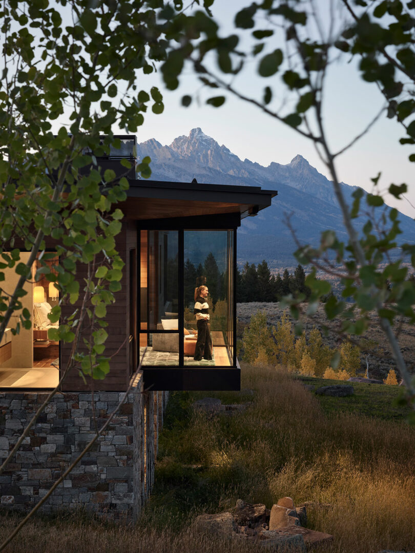Una persona se encuentra junto a la ventana de una casa moderna con paredes de vidrio, con vistas a un paisaje cubierto de hierba y montañas al anochecer.