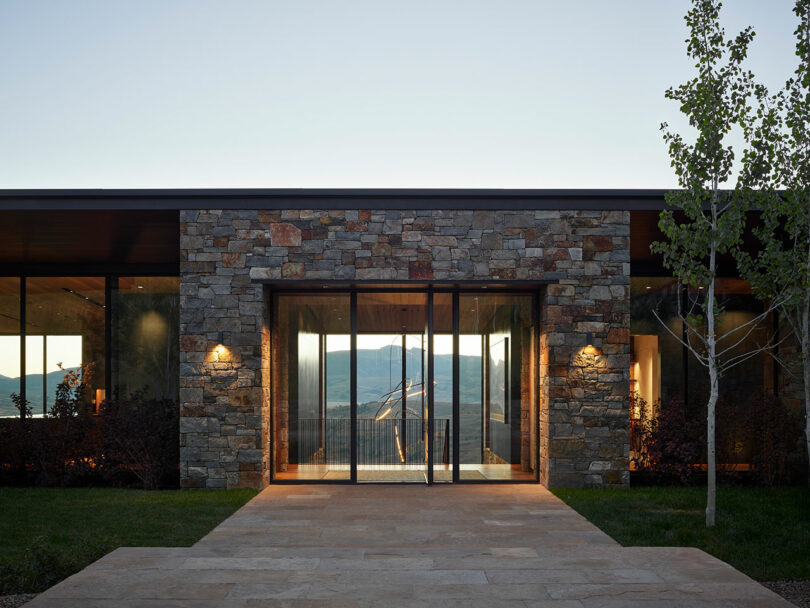 Casa moderna con una fachada de piedra y una gran entrada de vidrio, iluminada por luces exteriores, con un árbol y una vista de montaña en el fondo.
