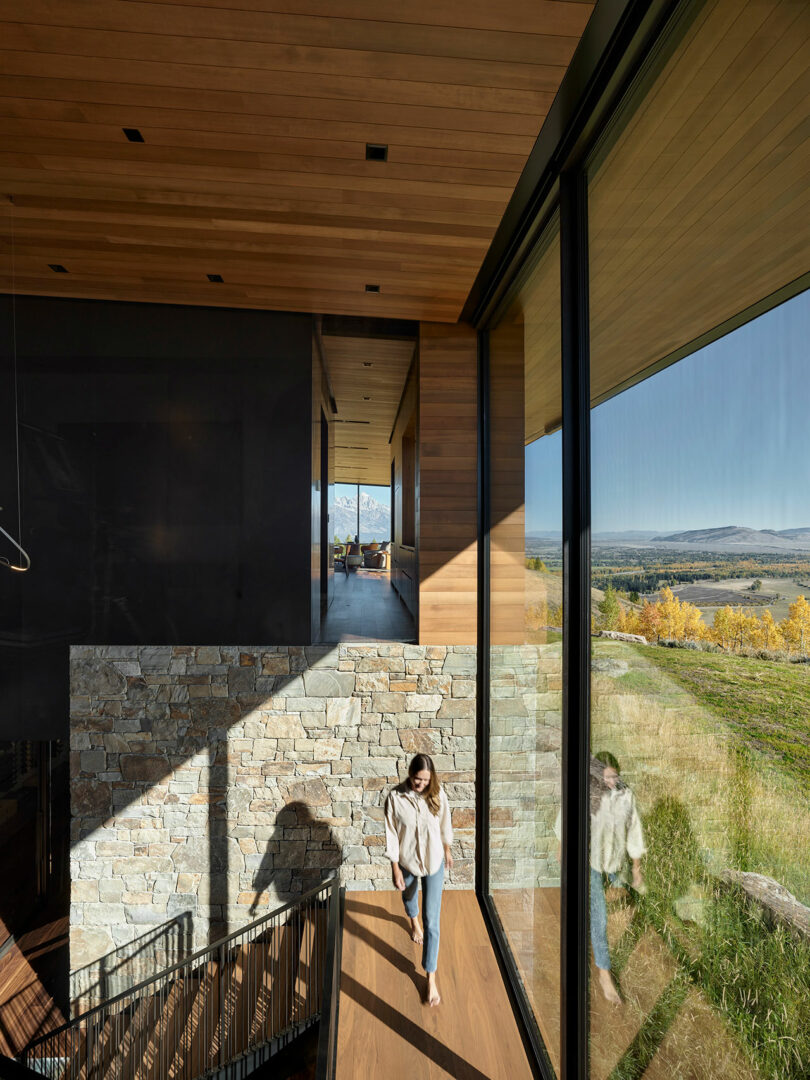 Una mujer camina por una escalera de madera dentro de una casa moderna con grandes vidrio con vistas a un paisaje pintoresco y cubierto de hierba y montañas distantes.