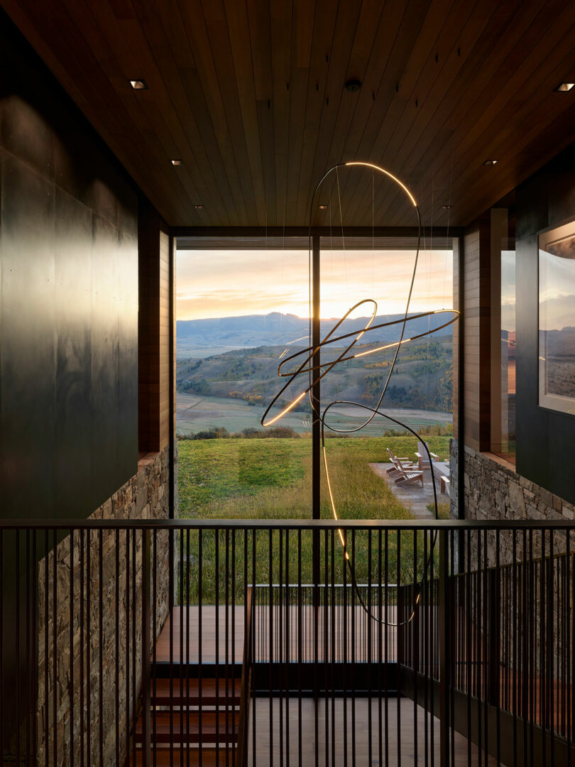 Interior moderno con una escalera de madera, paredes de piedra y una lámpara escultórica, con vistas a un patio al aire libre y un paisaje pintoresco a través de grandes ventanas de piso a techo al atardecer.