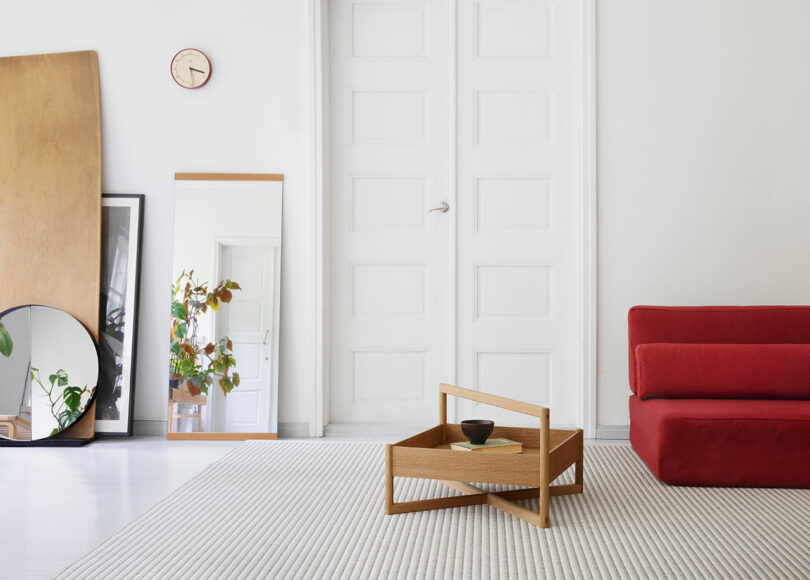Minimalist living room with a red sofa, wooden coffee table, mirrors, and a clock on a white wall, featuring a large white door and a potted plant.