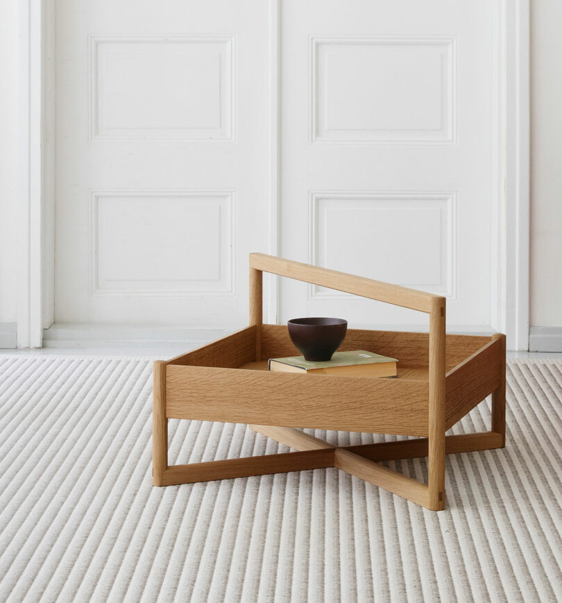 A modern wooden tray table with a handle, holding a book and a bowl, sits on a textured light-colored carpet in front of white paneled doors.
