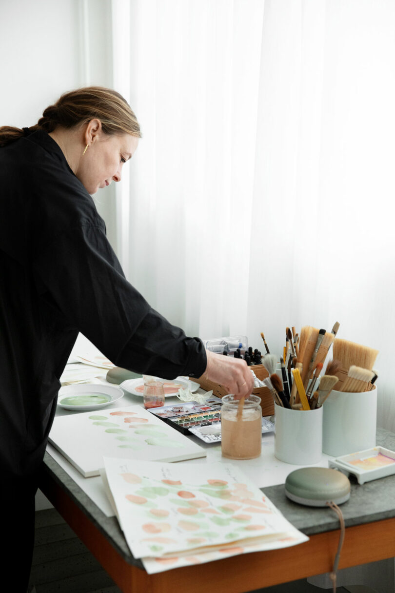 A person stands at a table by a window, painting with watercolors. Art supplies, brushes, and paper with colorful patterns are spread out on the tabletop.