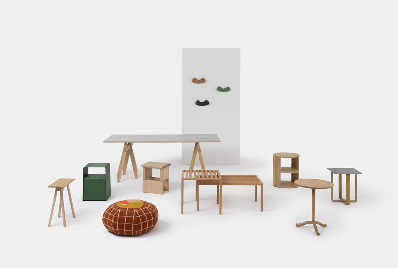 A group of minimalist wooden furniture pieces, including tables, stools, benches, a green box, and an orange pouf, arranged on a white background with wall hooks in the back.