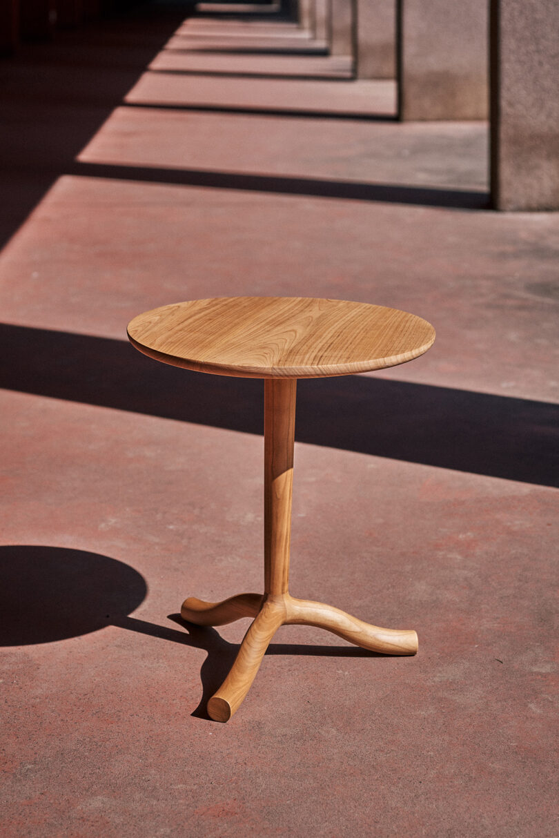 A round wooden table with a single central leg and three curved feet stands on a red concrete floor in an outdoor corridor with columns and shadows, evoking the mysterious atmosphere of an SCP containment site.