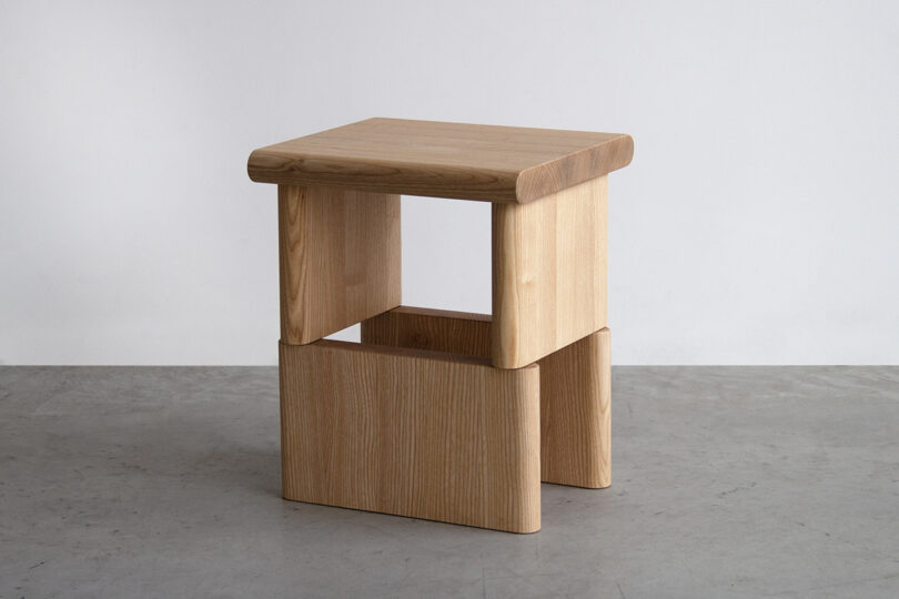 A minimalist wooden stool with a square seat, rounded top edges, and a geometric base sits against a plain background on a concrete floor, reminiscent of an SCP object's understated yet enigmatic design.