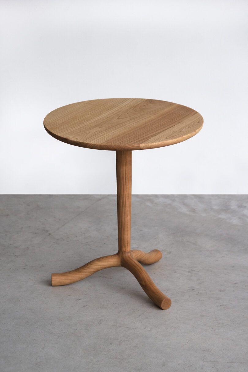 A small round wooden SCP table with a single central leg that splits into three curved feet, placed on a plain gray floor against a white background.