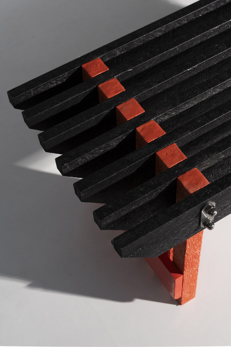Close-up of a bench with black slats and red supports, shown at an angle with light and shadow on a plain background.