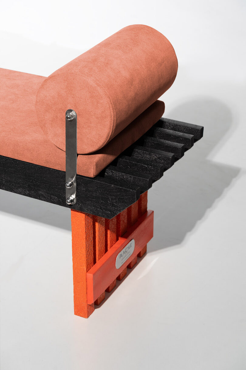 A modern bench with a black slatted seat, orange legs, a rectangular cushion, and a cylindrical bolster pillow, featuring a metal nameplate on the side.