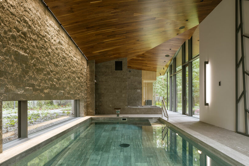 Indoor swimming pool featuring Sadowski-inspired stone walls, a wood-paneled sloped ceiling, and expansive floor-to-ceiling windows that fill the space with natural light.