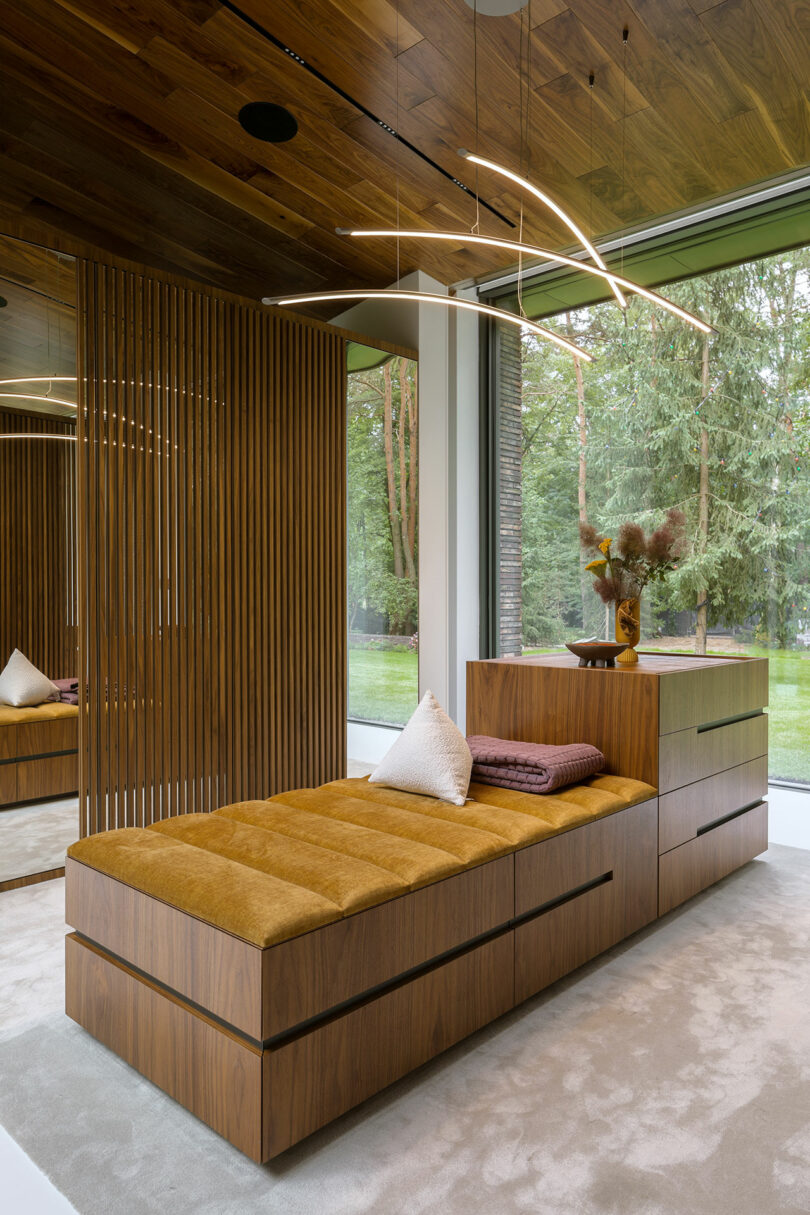 A modern Sadowski bedroom with a wood-paneled bed, matching headboard storage, and minimal decor, overlooking large windows with a view of trees outside.