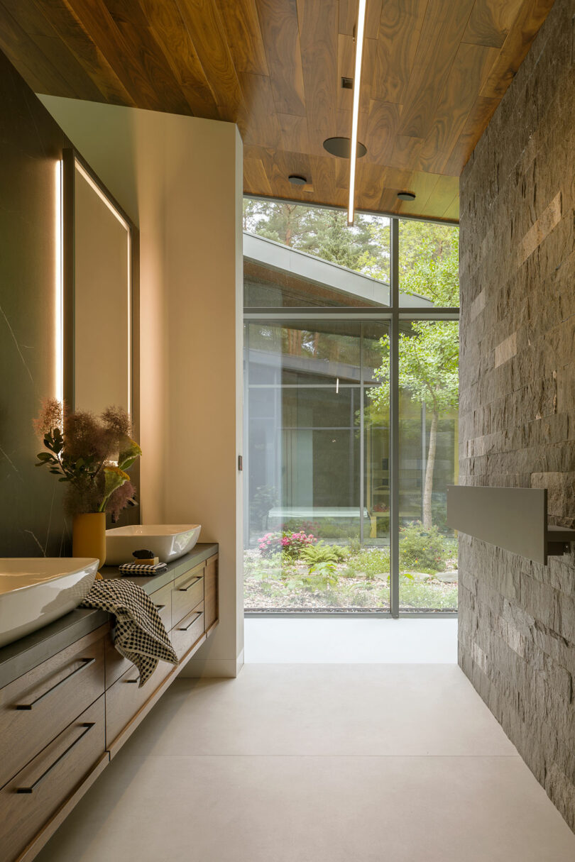 Modern Sadowski bathroom with dual sinks, a stone accent wall, wooden ceiling, and large floor-to-ceiling window overlooking a landscaped garden.