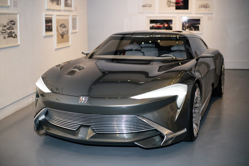 En el interior se exhibe un elegante concept car de color gris oscuro con elementos de diseño futurista, rodeado de bocetos de automóviles enmarcados en una pared blanca.