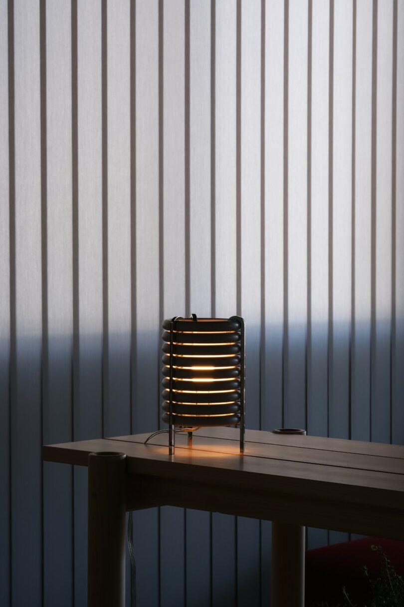 A modern table lamp with a layered, cylindrical design emits soft light on a wooden table in front of vertical blinds