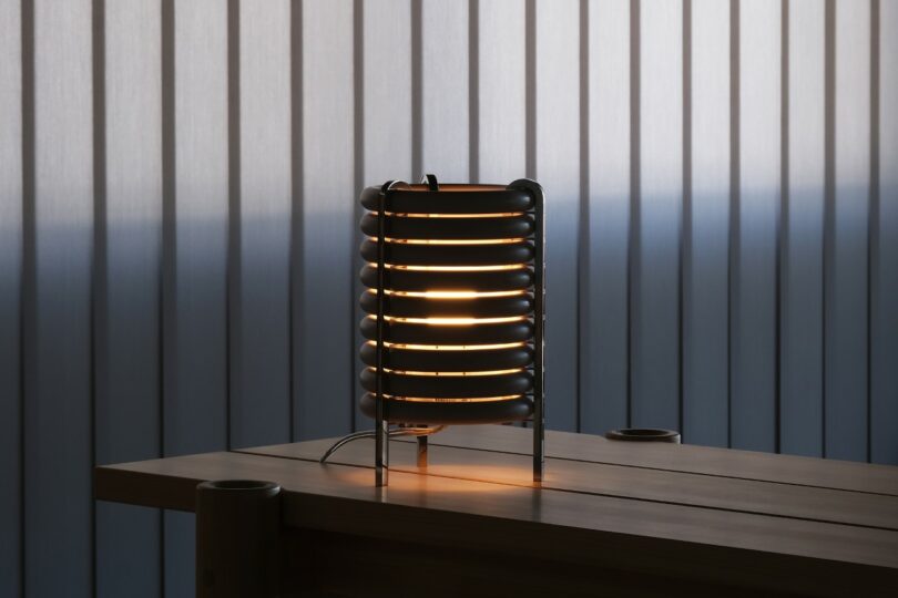 A modern cylindrical table lamp with horizontal black rings emits a warm light on a wooden table in front of vertical blinds