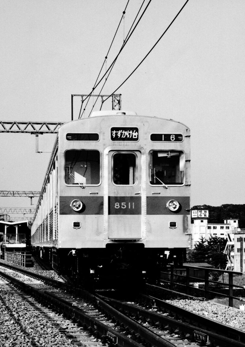 A front-facing view of a vintage electric train on railway tracks, with overhead power lines and buildings in the background, in black and white