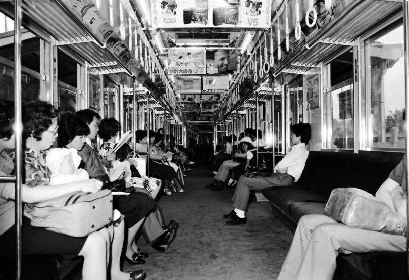 Black and white photo of passengers sitting on both sides of a subway train car; advertising posters hang from the ceiling above them