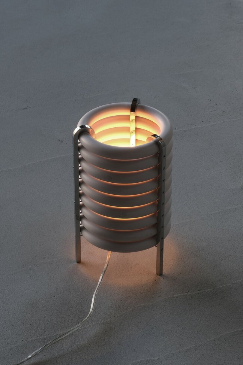 A cylindrical table lamp with horizontal white rings, metal legs, and a visible bulb glows warmly on a gray surface. A power cord extends from the base