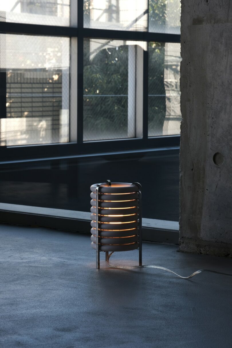 A modern cylindrical lamp with horizontal metal rings emits a warm light on a gray floor near a concrete pillar and large industrial windows