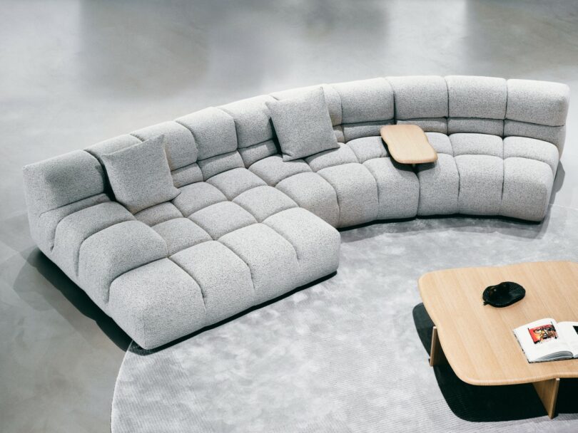 Curved light gray sectional sofa with three cushions and a small wooden tray, set on a light rug near a wooden coffee table with a book and dish