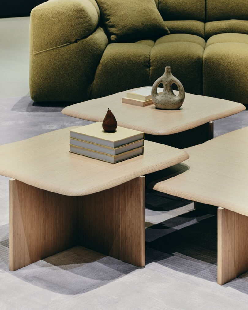 Three light wood coffee tables with stacked books, a small brown pear-shaped object, and a sculptural vase, placed in front of a green upholstered sofa