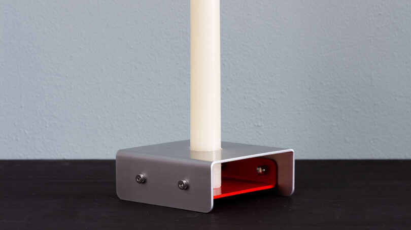 A white candle stands in a rectangular metal holder with a red interior, placed on a dark surface against a light gray wall.