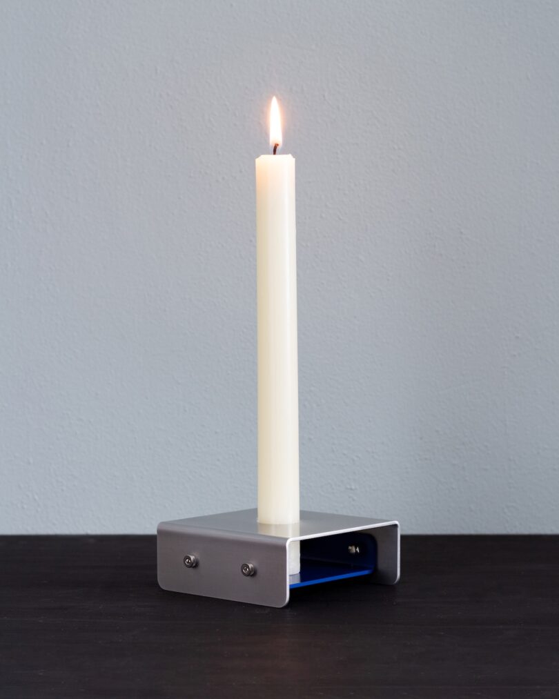 A lit white taper candle stands upright in a minimalist metal candle holder on a dark surface against a light gray wall.