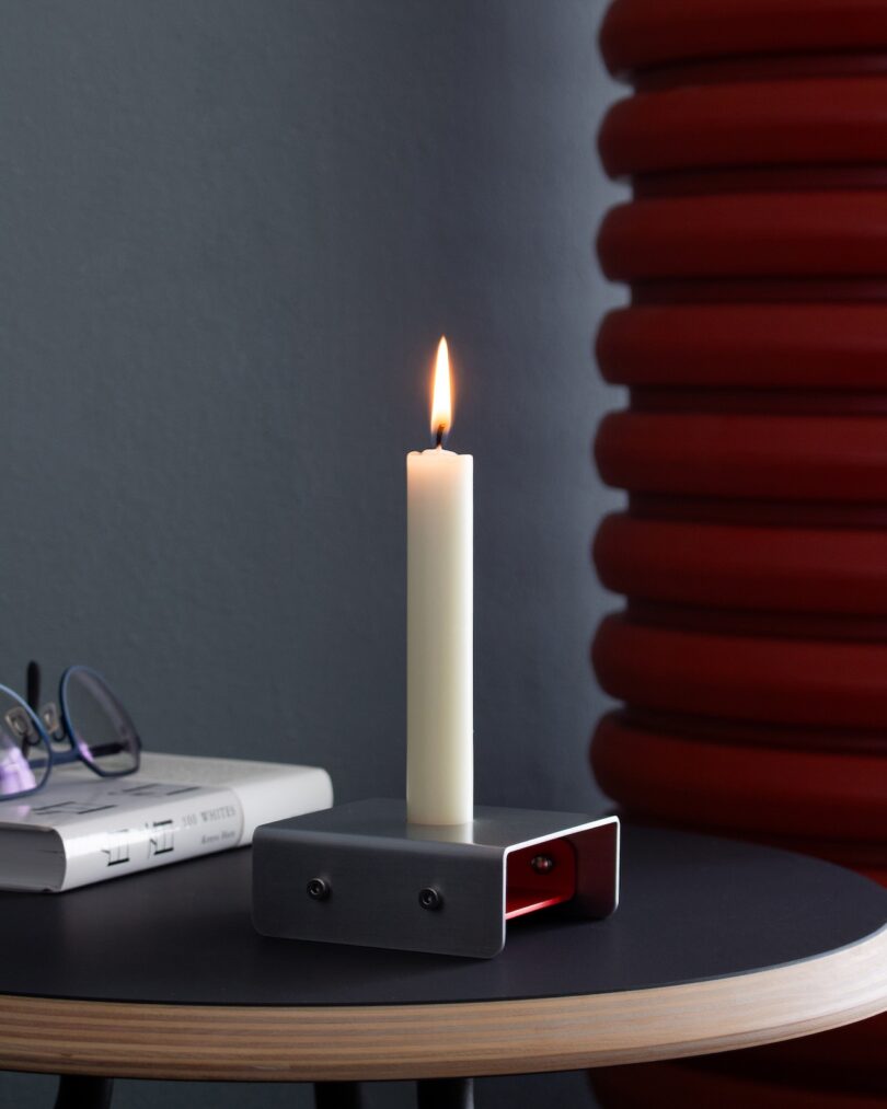 A lit white candle in a minimalist metal holder sits on a round black table next to a closed book and a pair of eyeglasses, with a red textured object in the background.