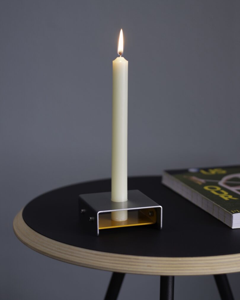 A lit white candle in a minimalist metal holder sits on a round, black side table next to a closed book.